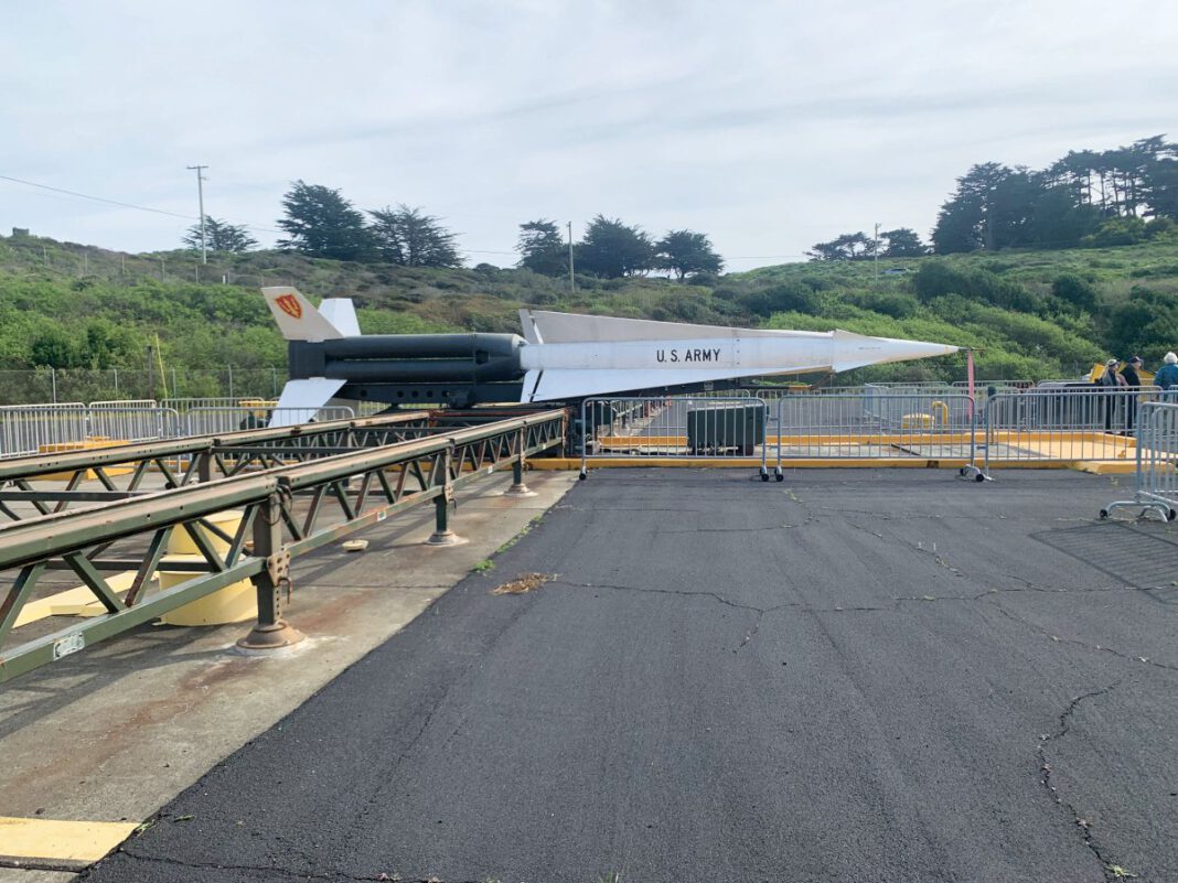 A long white missile stands horizontally on a metal launch platform beside trees and hills at a former military installation.