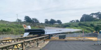 A long white missile stands horizontally on a metal launch platform beside trees and hills at a former military installation.