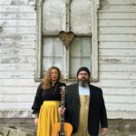 Around the Sun and Up the Wall Two musicians standing with an acoustic guitar in front of a weathered white building