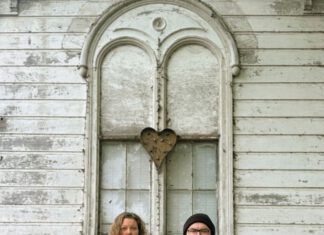Two musicians standing with an acoustic guitar in front of a weathered white building