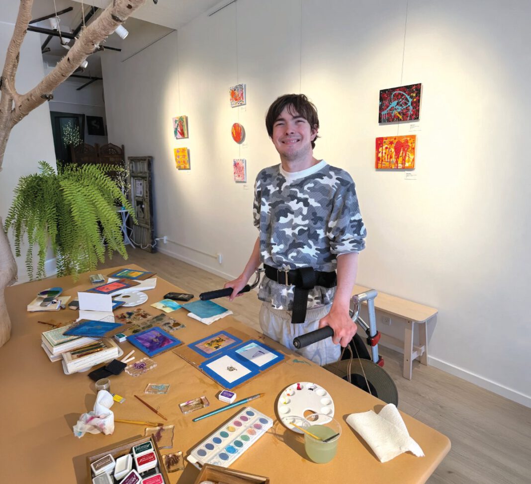 Artist standing beside a table covered with paintings and art supplies in a gallery