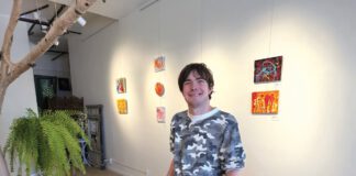 Artist standing beside a table covered with paintings and art supplies in a gallery