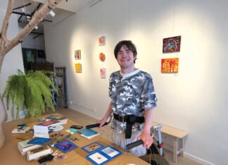 Sausalito Art Collective, Ancestral Land and Fiesta Forever Artist standing beside a table covered with paintings and art supplies in a gallery