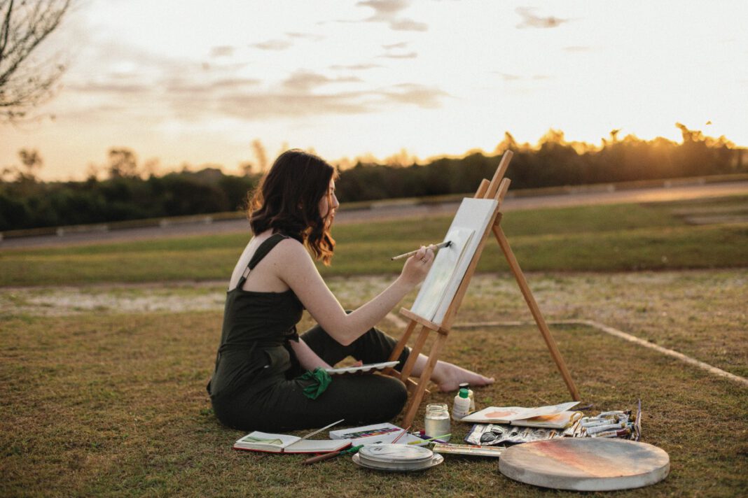 Artist painting on a canvas outdoors beside an easel at sunset
