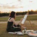 Artist painting on a canvas outdoors beside an easel at sunset