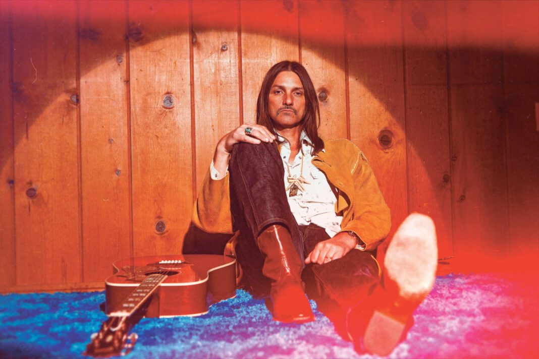 Duane Betts sits on a blue carpet next to a guitar.
