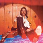 Duane Betts sits on a blue carpet next to a guitar.