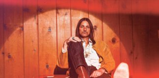 Duane Betts sits on a blue carpet next to a guitar.