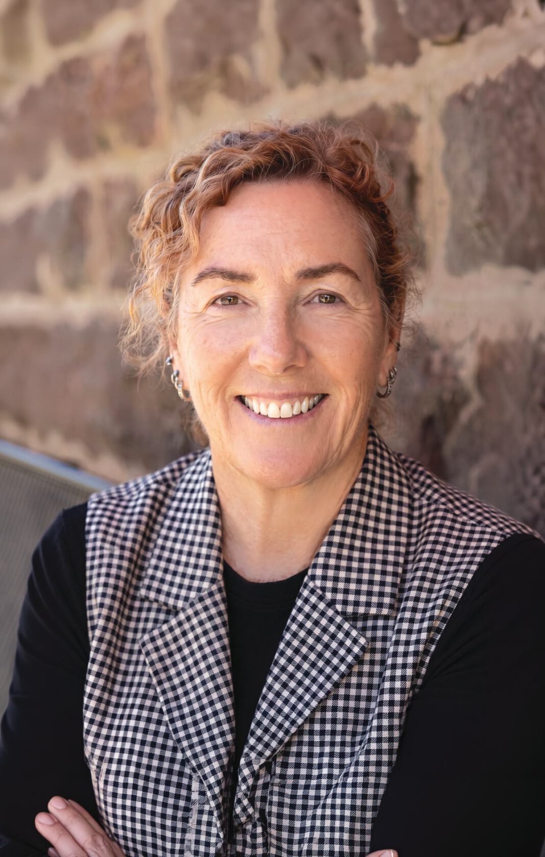 A smiling woman with her hair pulled back wears a black long-sleeve shirt and a black-and-white patterned vest.