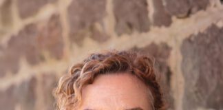 A smiling woman with her hair pulled back wears a black long-sleeve shirt and a black-and-white patterned vest.