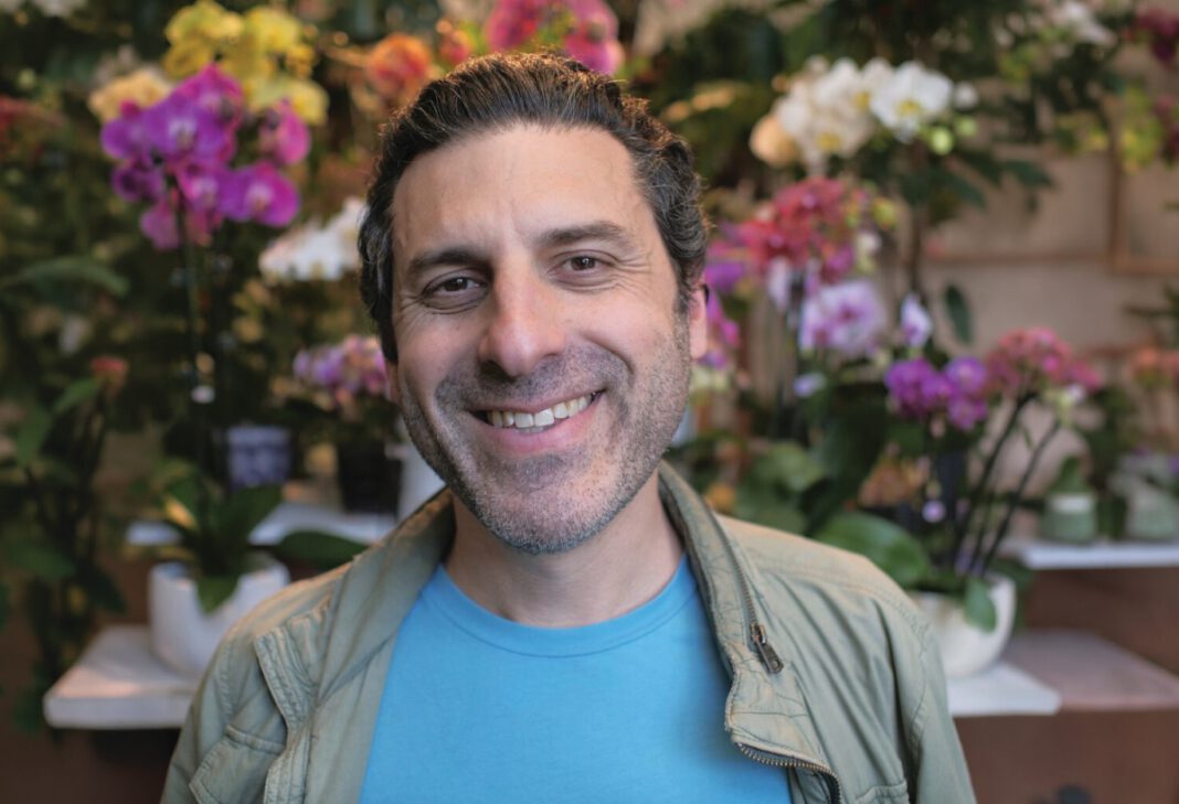 A smiling man in a light jacket stands in front of colorful orchids and other flowers.