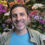 A smiling man in a light jacket stands in front of colorful orchids and other flowers.
