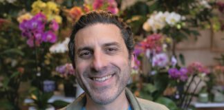 A smiling man in a light jacket stands in front of colorful orchids and other flowers.