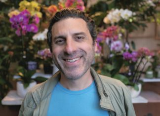 A smiling man in a light jacket stands in front of colorful orchids and other flowers.
