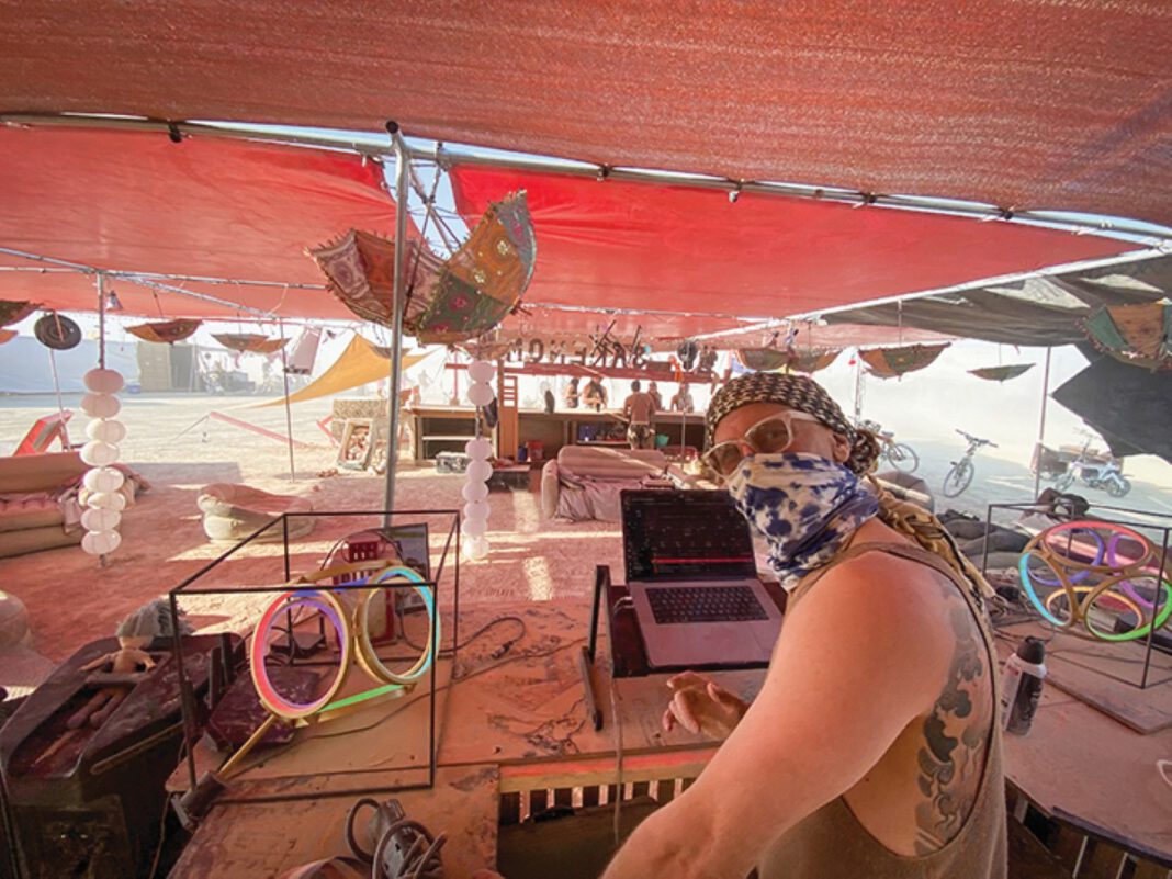 A man in sunglasses and a bandana looks toward the camera while working at a laptop under a red shade canopy. Glowing LED rings and bicycles are nearby.