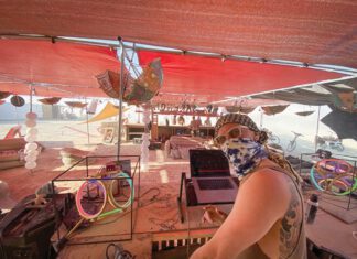 Burning Man Radio, KSVY’s RJ Grooms Keeps the Signal Hot A man in sunglasses and a bandana looks toward the camera while working at a laptop under a red shade canopy. Glowing LED rings and bicycles are nearby.
