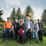 Six musicians stand together outdoors near pine trees at dusk.