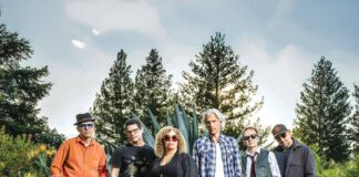 Six musicians stand together outdoors near pine trees at dusk.