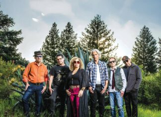 Six musicians stand together outdoors near pine trees at dusk.