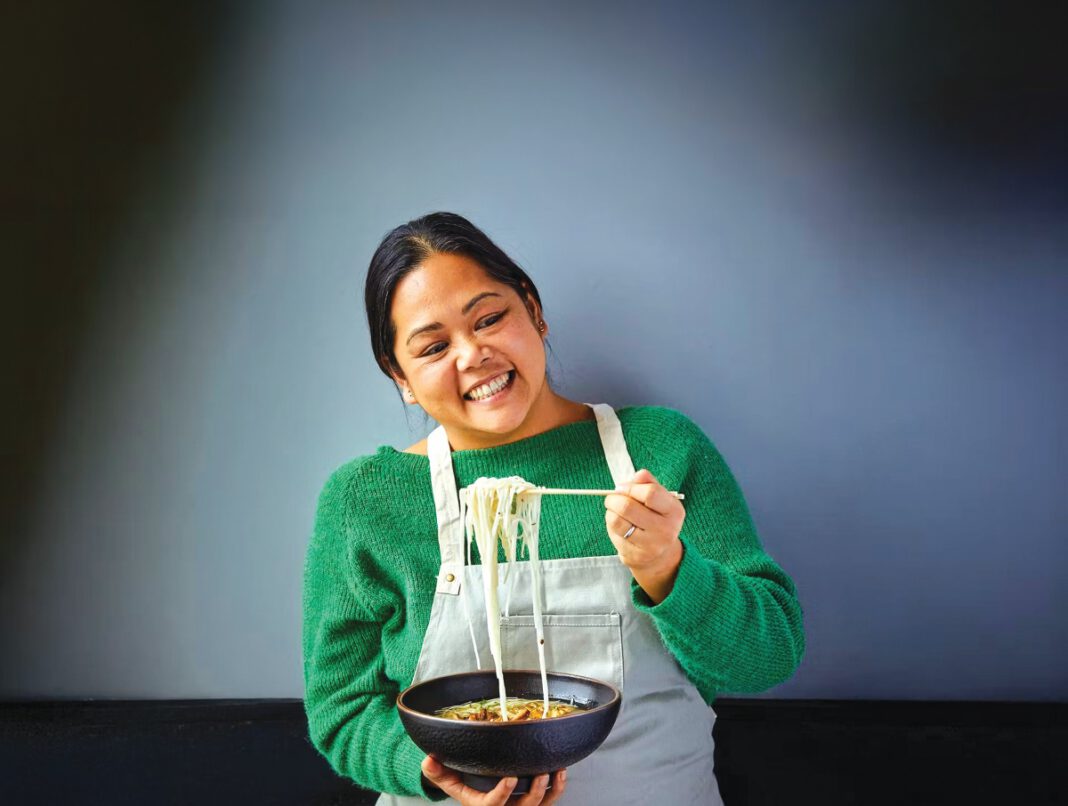 A smiling woman in a green sweater and apron lifts noodles with chopsticks over a bowl of soup.