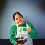 A smiling woman in a green sweater and apron lifts noodles with chopsticks over a bowl of soup.