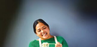 A smiling woman in a green sweater and apron lifts noodles with chopsticks over a bowl of soup.