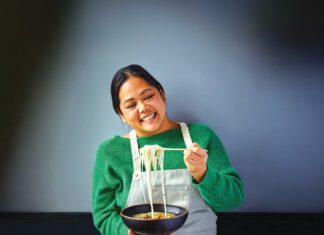A smiling woman in a green sweater and apron lifts noodles with chopsticks over a bowl of soup.