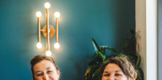 Two smiling women hold fancy frosted cakes on glass pedestal stands.