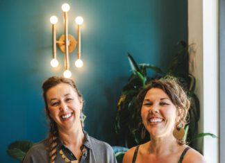 The Scoop, Sisterhood of Nimble & Finn’s Two smiling women hold fancy frosted cakes on glass pedestal stands.