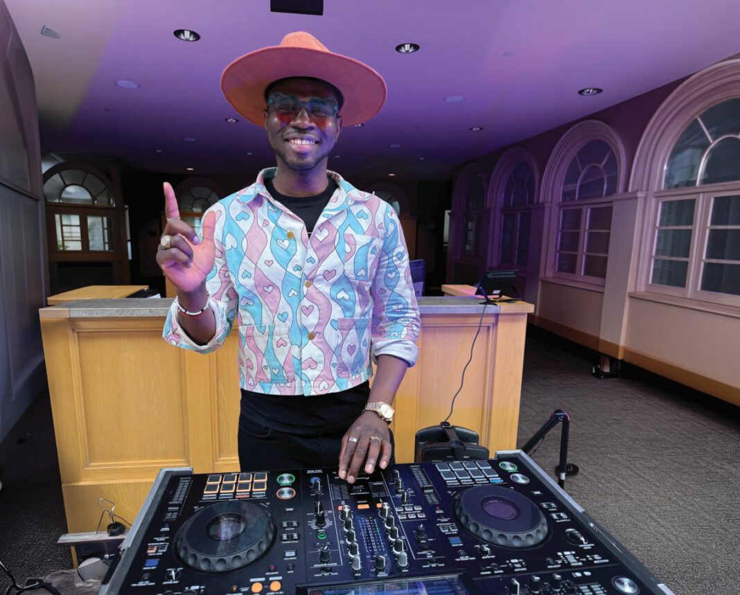 A DJ in a wide-brimmed pink hat and heart-patterned jacket extends his index finger up while working a turntable in a room with purple light.