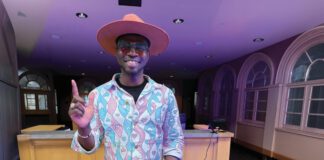 A DJ in a wide-brimmed pink hat and heart-patterned jacket extends his index finger up while working a turntable in a room with purple light.