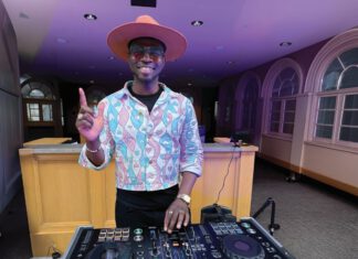 AfroBrasil, Acorns & Altered States A DJ in a wide-brimmed pink hat and heart-patterned jacket extends his index finger up while working a turntable in a room with purple light.