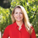She Has an Eye for Wine: Rebecka Deike’s Winemaking Path A woman with long blonde hair and a red button-down shirt smiles in front of green grapevines.