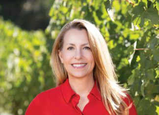 A woman with long blonde hair and a red button-down shirt smiles in front of green grapevines.
