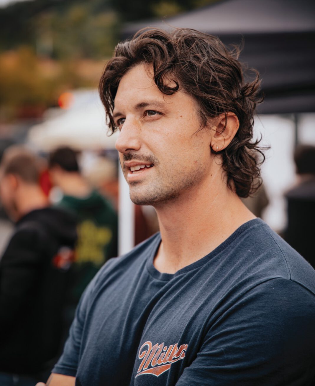 A man with curly brown hair and a mustache wears a navy t-shirt.