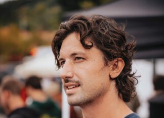 The Happys’ Guitarist Pedals Biketoberfest Into Its 20th Year A man with curly brown hair and a mustache wears a navy t-shirt.