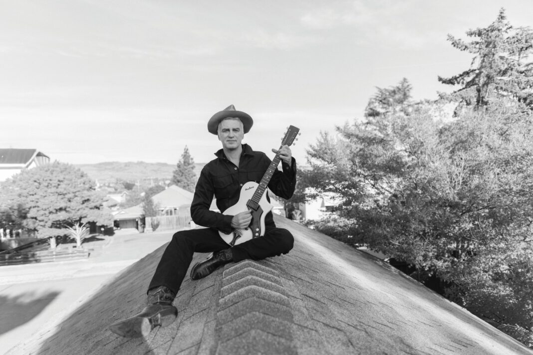 A black-and-white photo of a man in a wide-brimmed hat and dark clothing sitting cross-legged on a rooftop peak, holding a guitar.