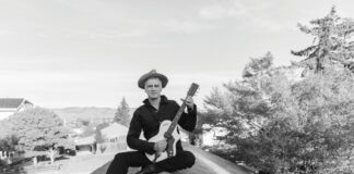 A black-and-white photo of a man in a wide-brimmed hat and dark clothing sitting cross-legged on a rooftop peak, holding a guitar.