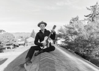 Brighter Still: 10th Time’s the Charm for Sean Hayes A black-and-white photo of a man in a wide-brimmed hat and dark clothing sitting cross-legged on a rooftop peak, holding a guitar.