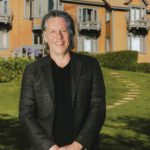 A smiling man in a blazer stands on a lawn in front of a large shingled house with balconies.