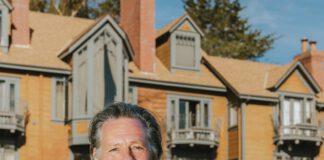 A smiling man in a blazer stands on a lawn in front of a large shingled house with balconies.