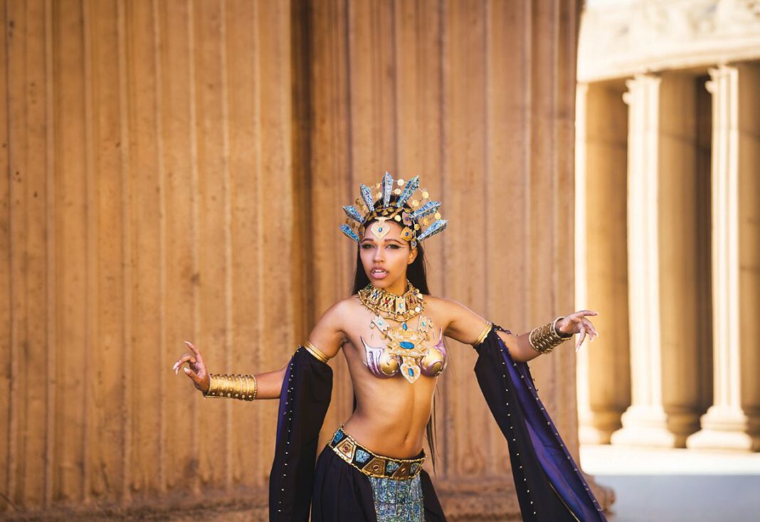 A cosplayer in an ornate gold headdress and jeweled costume poses dramatically in front of stone columns.