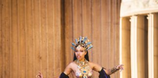 A cosplayer in an ornate gold headdress and jeweled costume poses dramatically in front of stone columns.