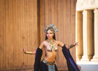A cosplayer in an ornate gold headdress and jeweled costume poses dramatically in front of stone columns.