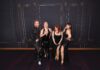 Plants, Prost & Perfect Harmony Four performers in glittering black outfits pose together against a dark, ornate wallpapered backdrop.