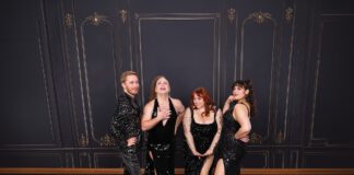 Four performers in glittering black outfits pose together against a dark, ornate wallpapered backdrop.