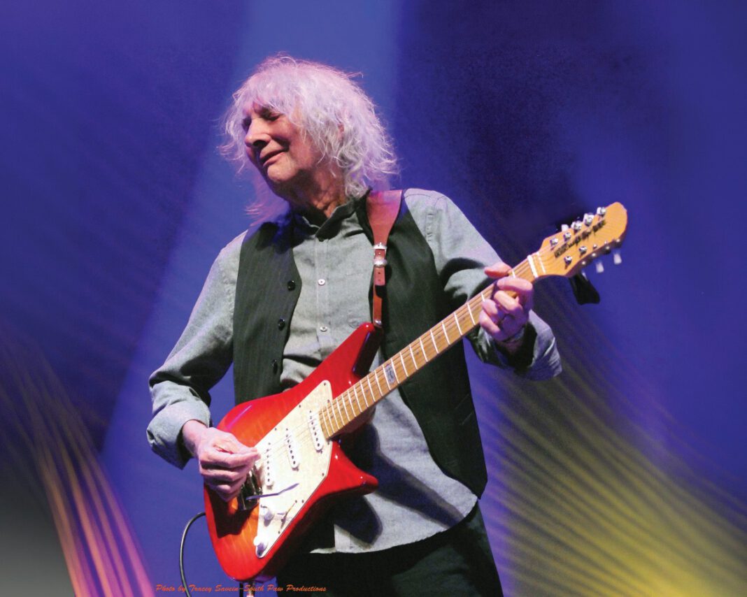 Albert Lee, 82, with light, wavy hair plays a red electric guitar onstage under blue and gold lighting.