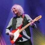 Albert Lee, 82, with light, wavy hair plays a red electric guitar onstage under blue and gold lighting.