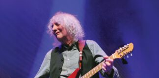 Albert Lee, 82, with light, wavy hair plays a red electric guitar onstage under blue and gold lighting.
