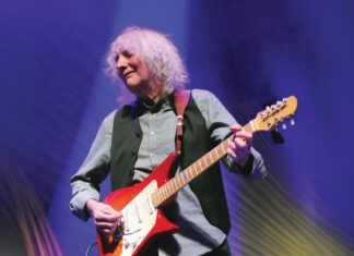 Albert Lee, 82, with light, wavy hair plays a red electric guitar onstage under blue and gold lighting.